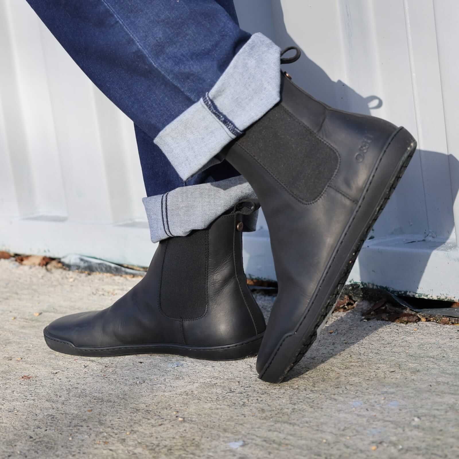 Person wearing black leather boots with rolled-up jeans on a concrete surface.