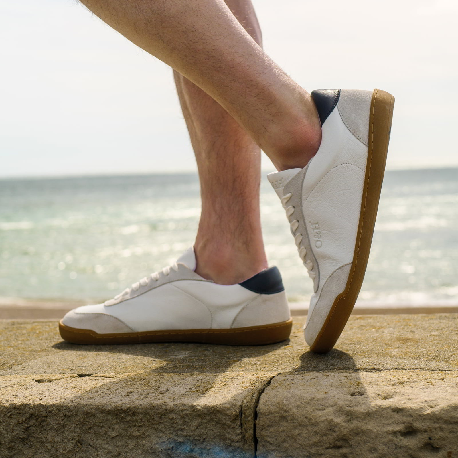 model standing in aspen white barefoot trainers with shorts on in a beach setting