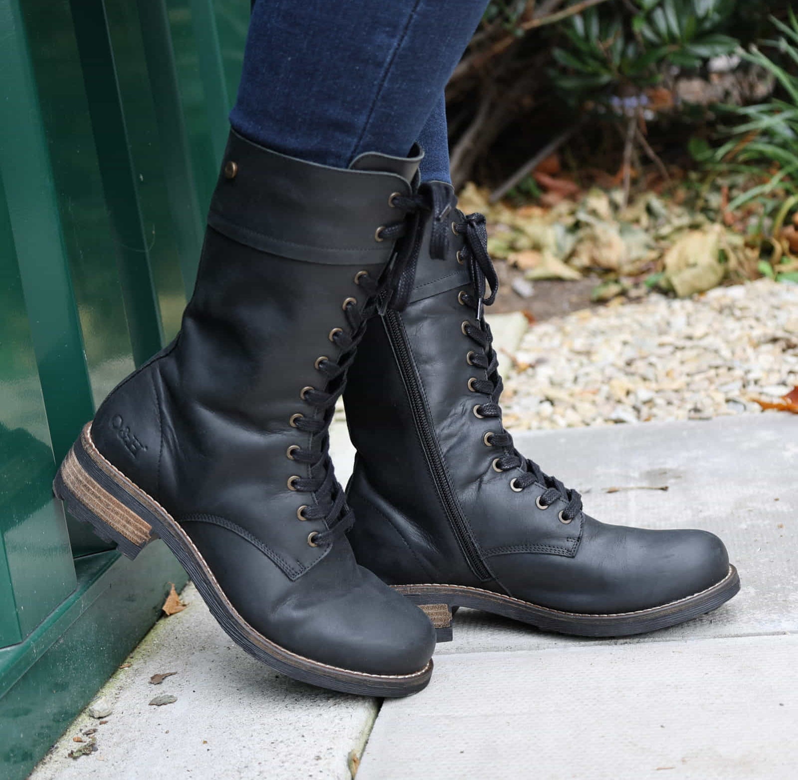 Black Bridge 12 military boots worn with blue jeans outdoors, side view against a green wall