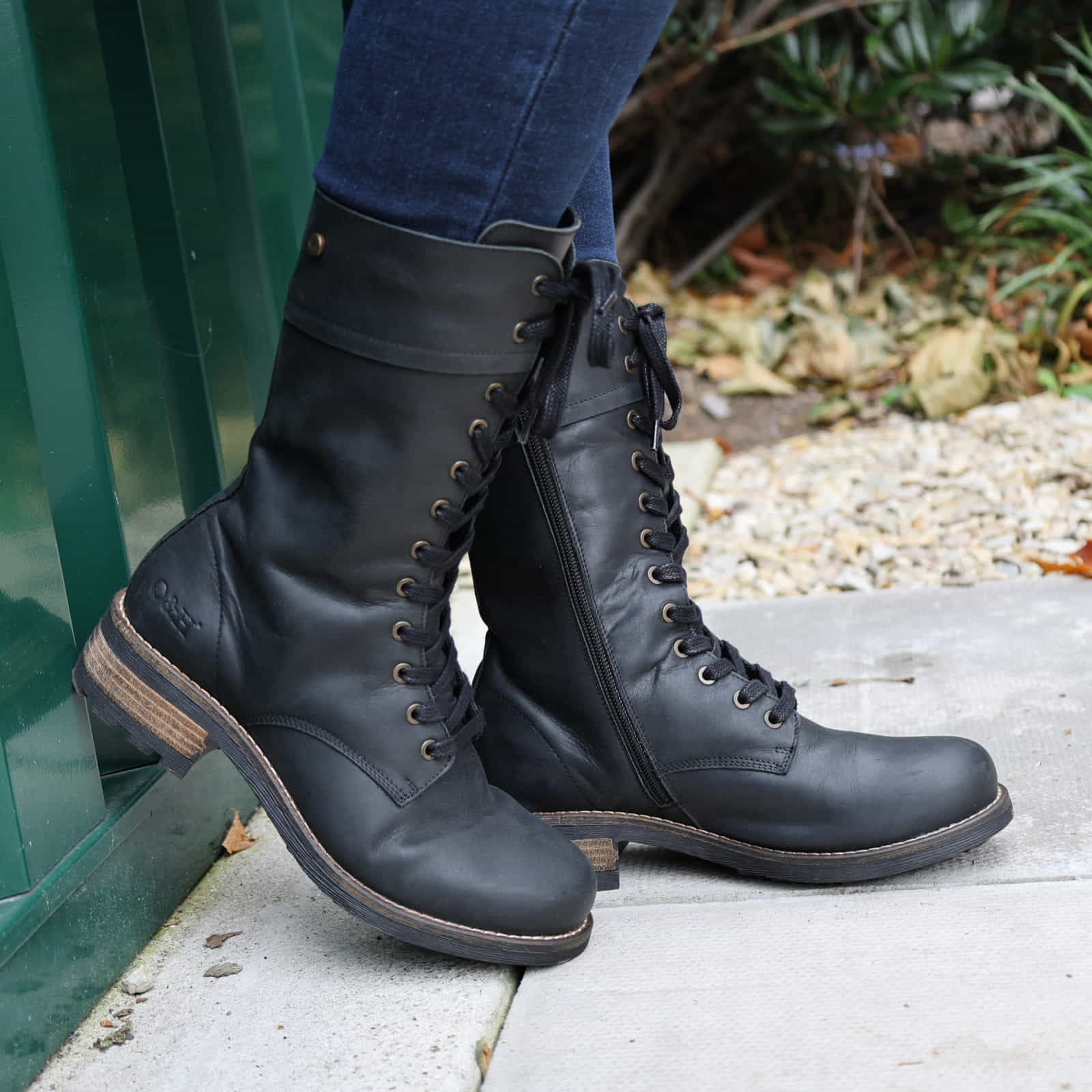 Black Bridge 12 military boots worn with blue jeans outdoors, side view against a green wall