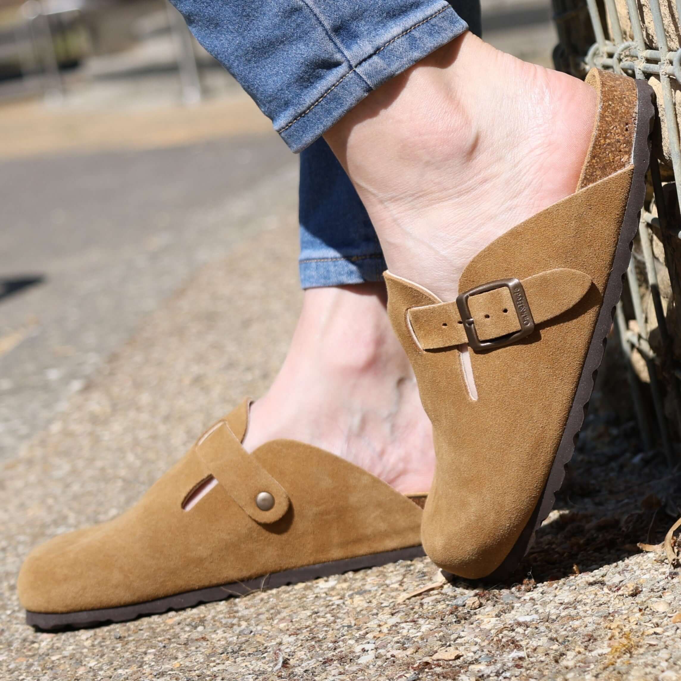 model wearing tan gibraltar clogs with blue jeans leaning against a wall