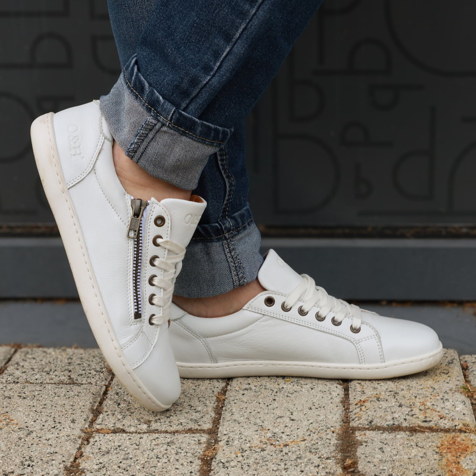 model standing outdoors wearing hazel zip barefoot trainers in white with blue jeans