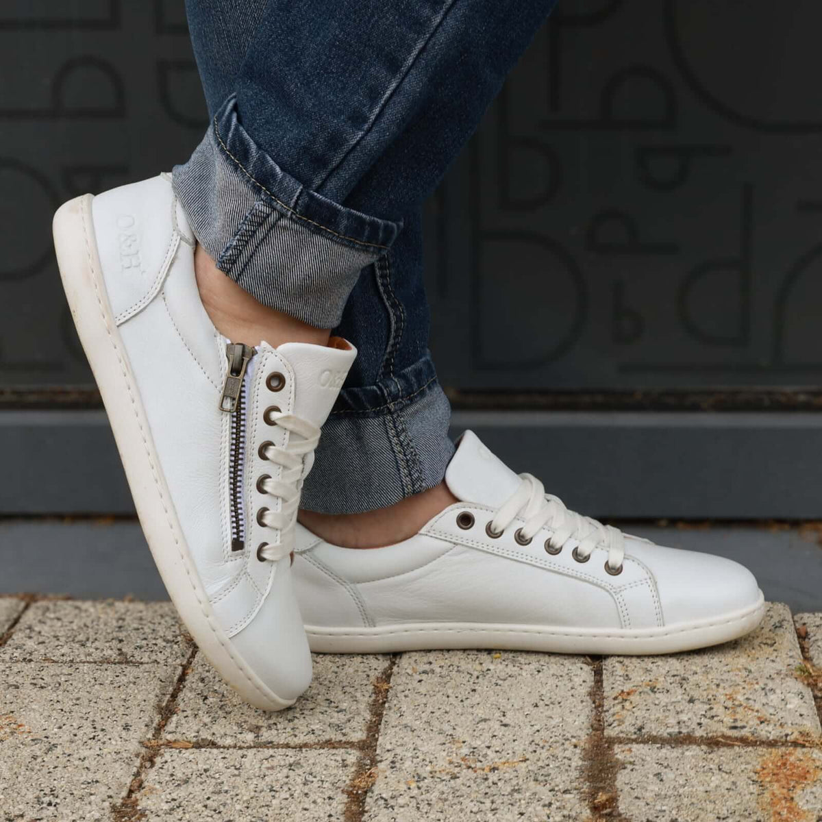 model standing outdoors wearing hazel zip barefoot trainers in white with blue jeans
