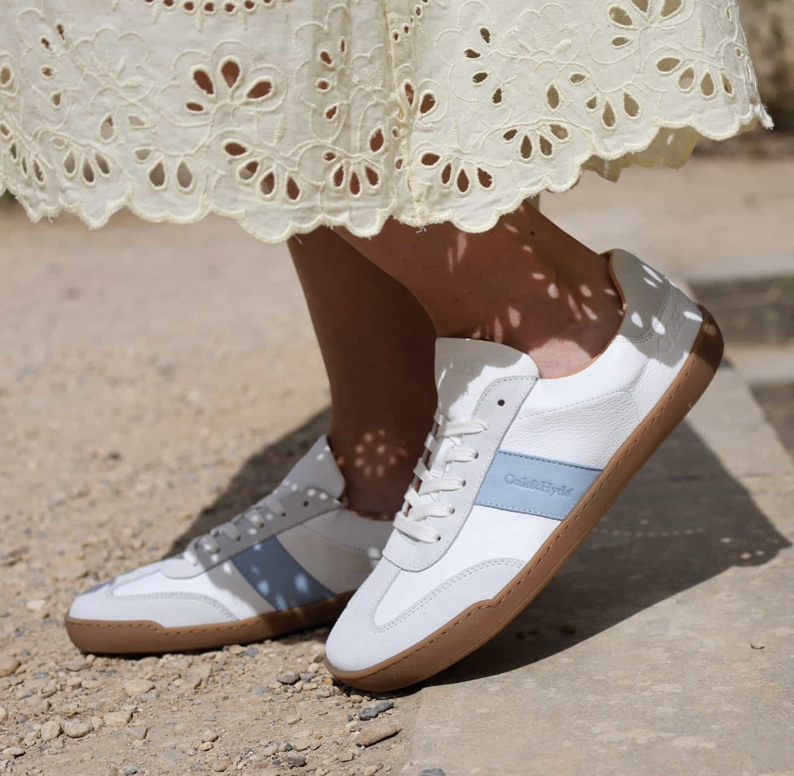 model wearing white barefoot trainers showing Oak & Hyde branding and blue stripe on gravel pavement
