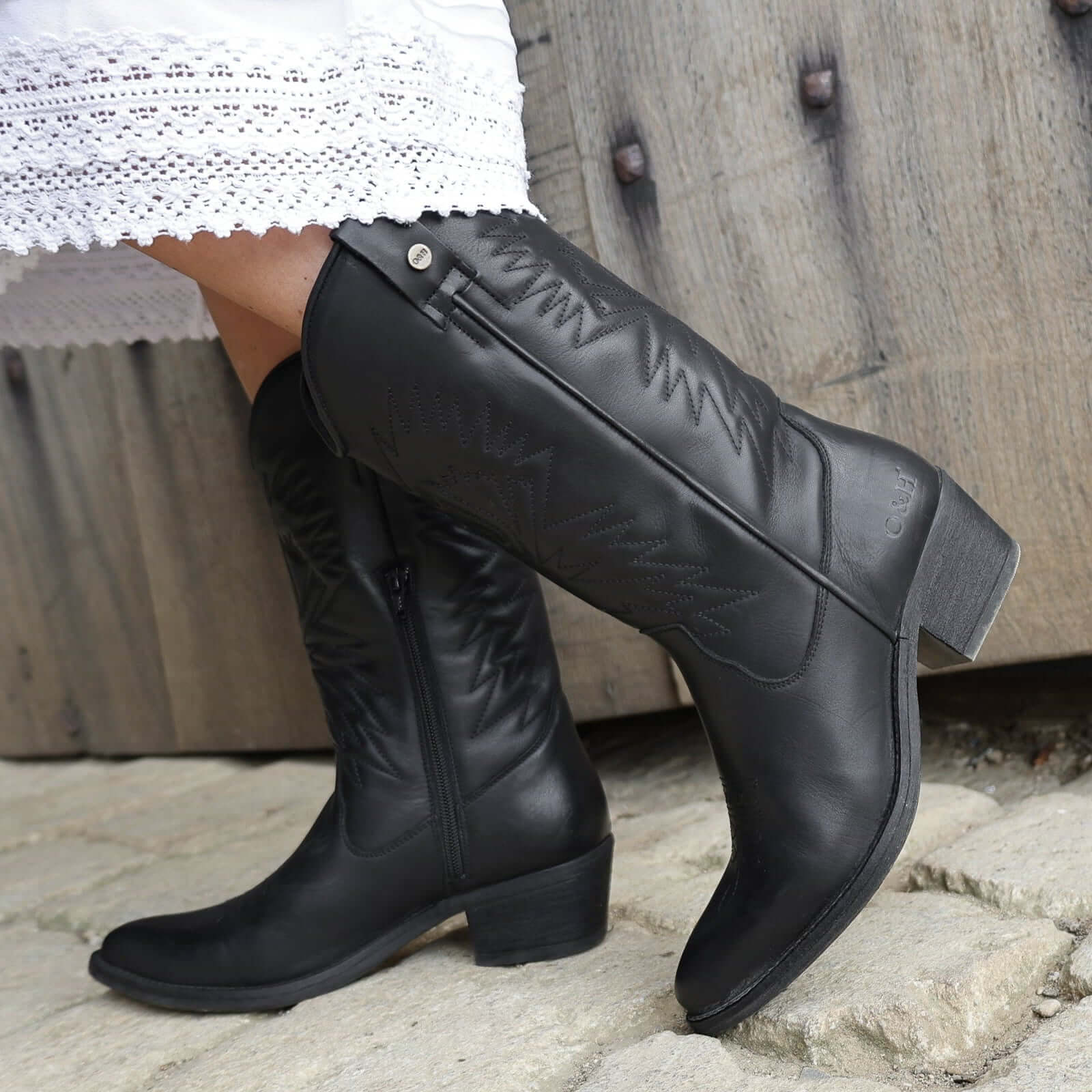 model wearing black leather texan hi cowboy boots with a white dress on a stone floor