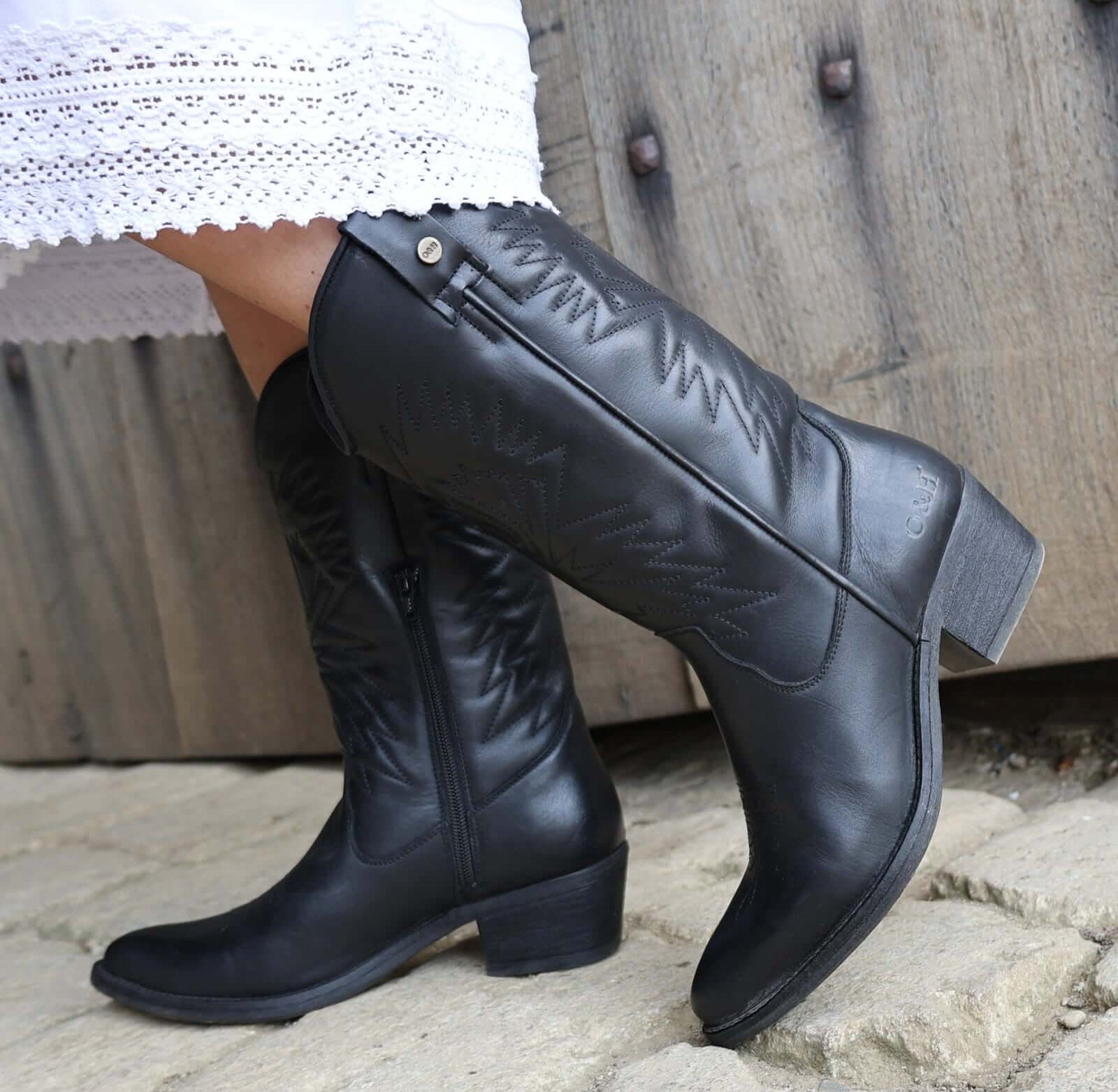 model wearing black leather texan hi cowboy boots with a white dress on a stone floor