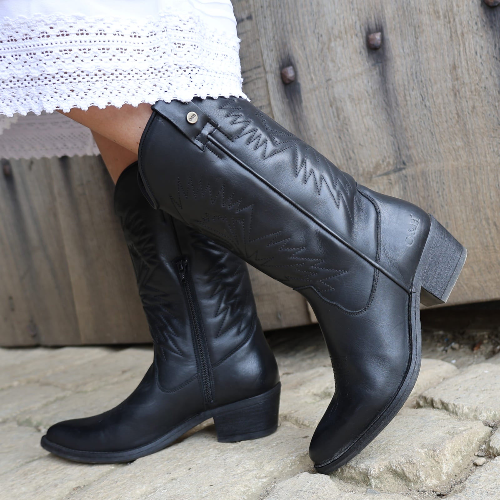 model wearing black leather texan hi cowboy boots with a white dress on a stone floor