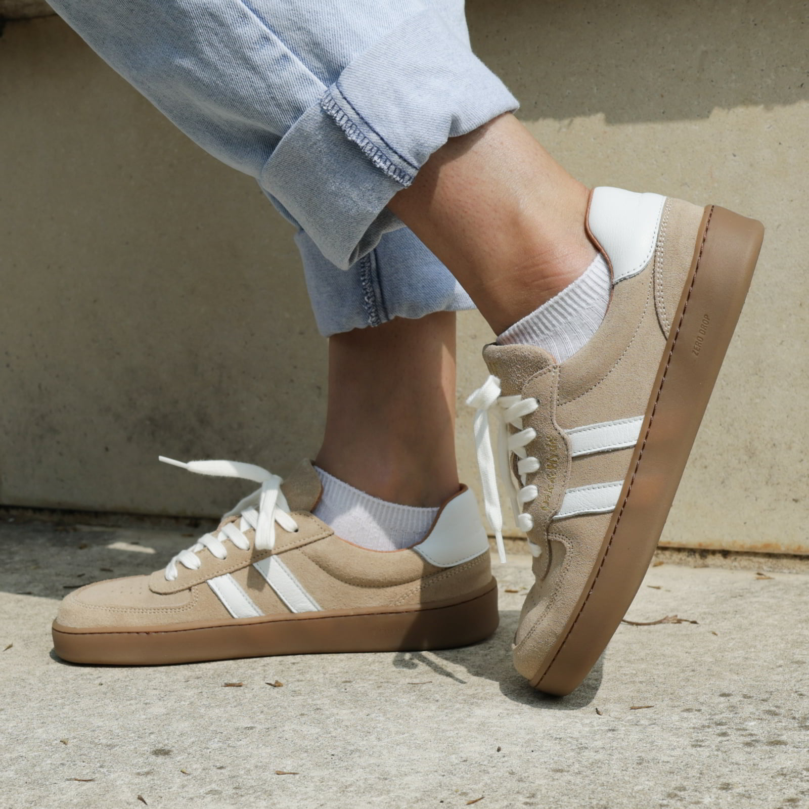 model standing outdoors wearing womens larch barefoot trainers in beige with blue jeans
