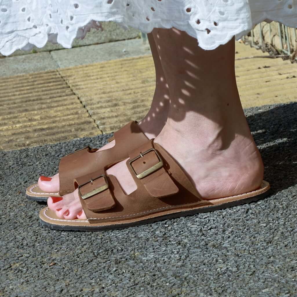 model standing outdoors with acacia womens leather barefoot sandals in tan being worn with a white dress