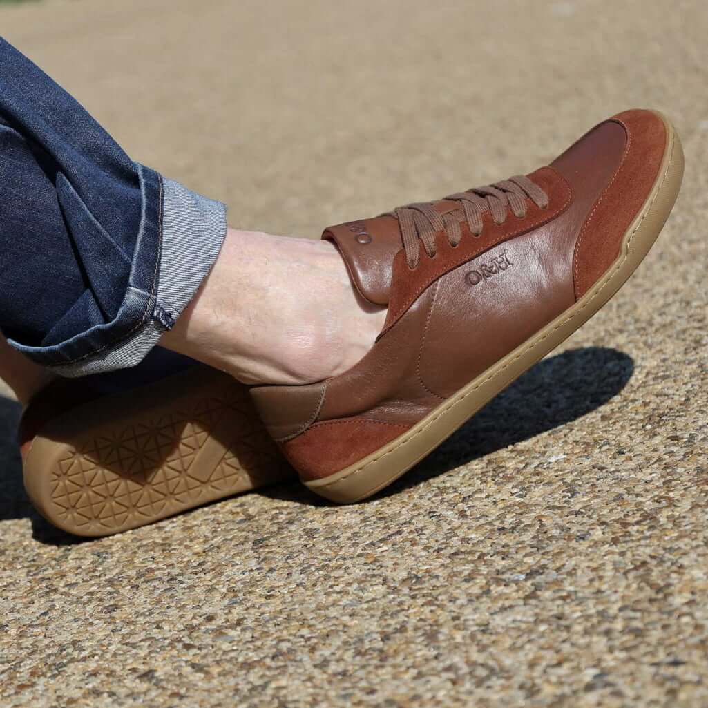 model wearing aspen barefoot trainers in tan with blue jeans sitting on bench