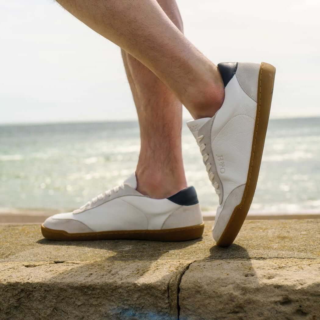 model standing in aspen white barefoot trainers with shorts on in a beach setting