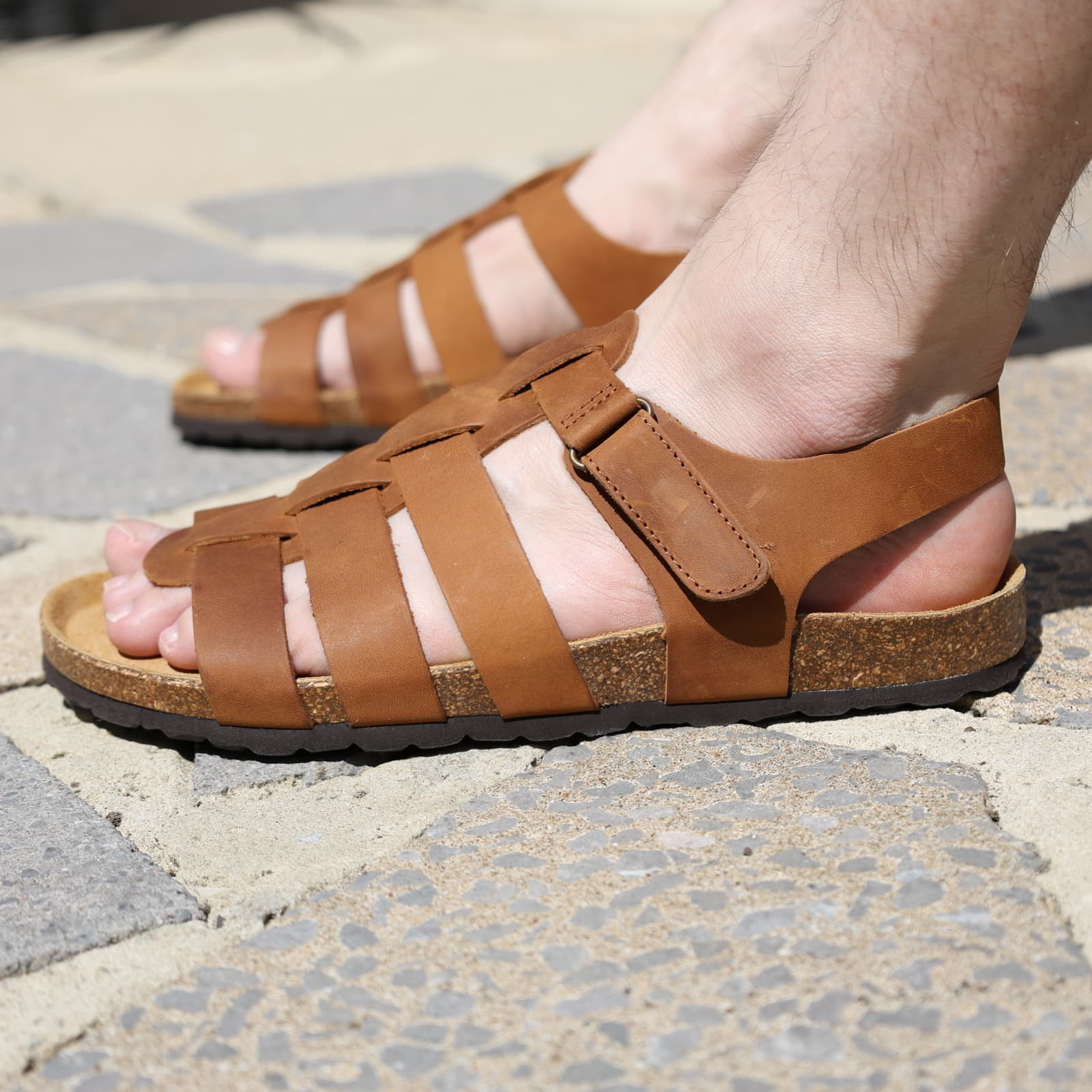Tan leather fisherman sandals worn on foot while standing on sunlit stone paving outdoors