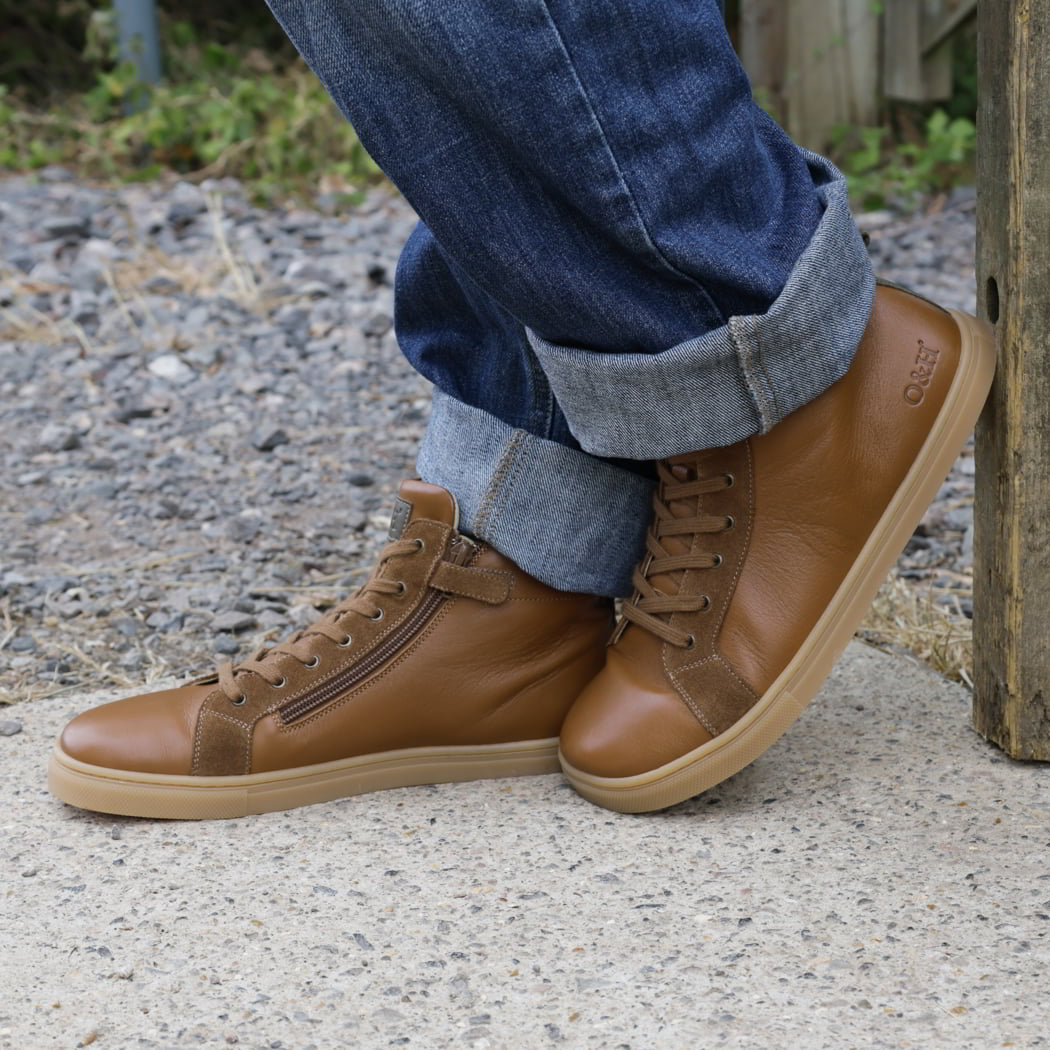 person wearing mens elm barefoot boots with blue jeans leaning against a gate