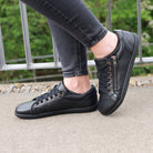 model standing outdoors wearing hazel zip barefoot trainers in black