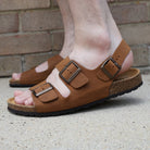 Tan leather fisherman sandals worn on foot, standing on concrete ground in front of a brick wall