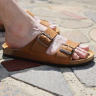 Person wearing tan leather fisherman sandals with buckle straps on outdoor stone paving