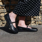 person wearing olive barefoot mary jane shoes in black with black spotty trousers standing outside