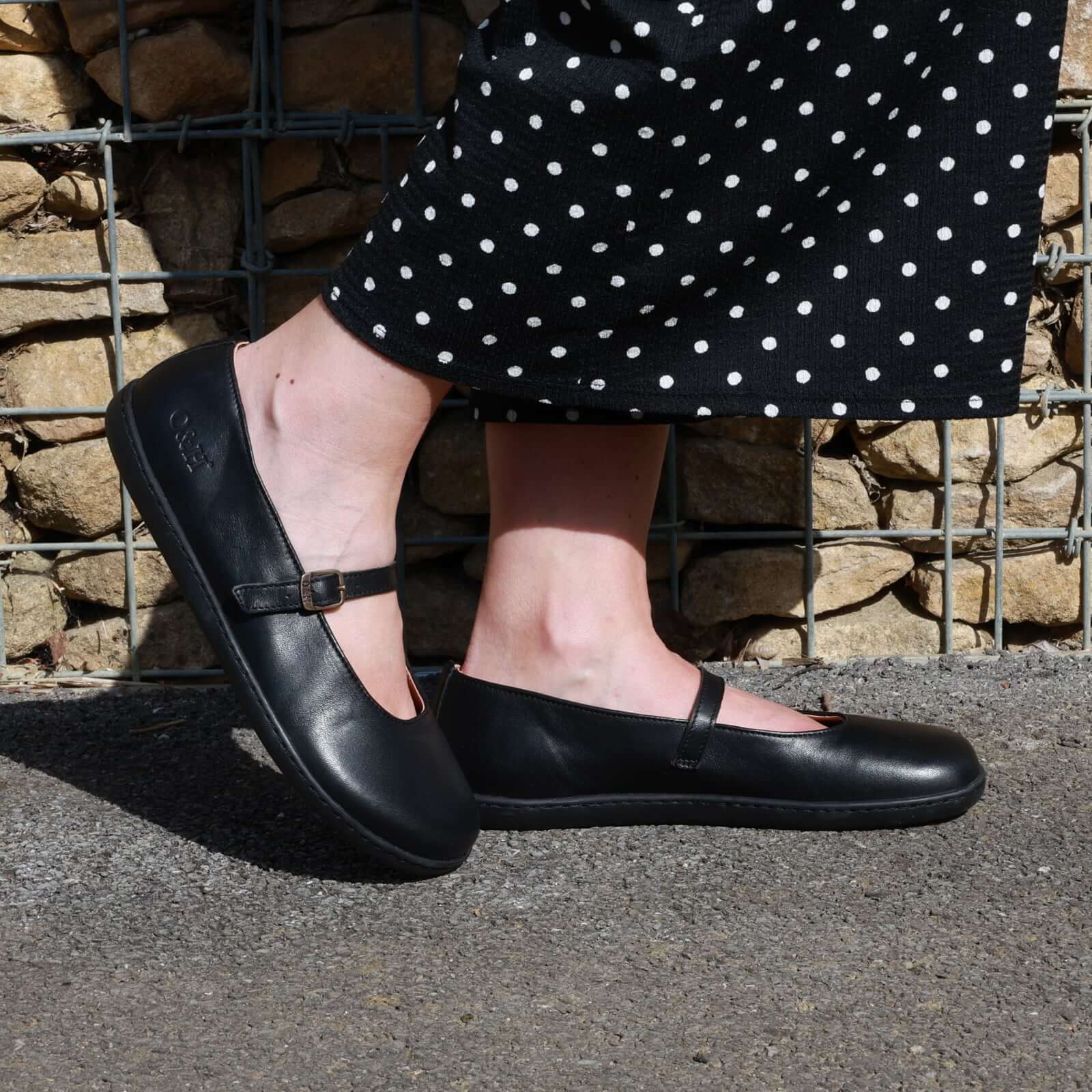 person wearing olive barefoot mary jane shoes in black with black spotty trousers standing outside