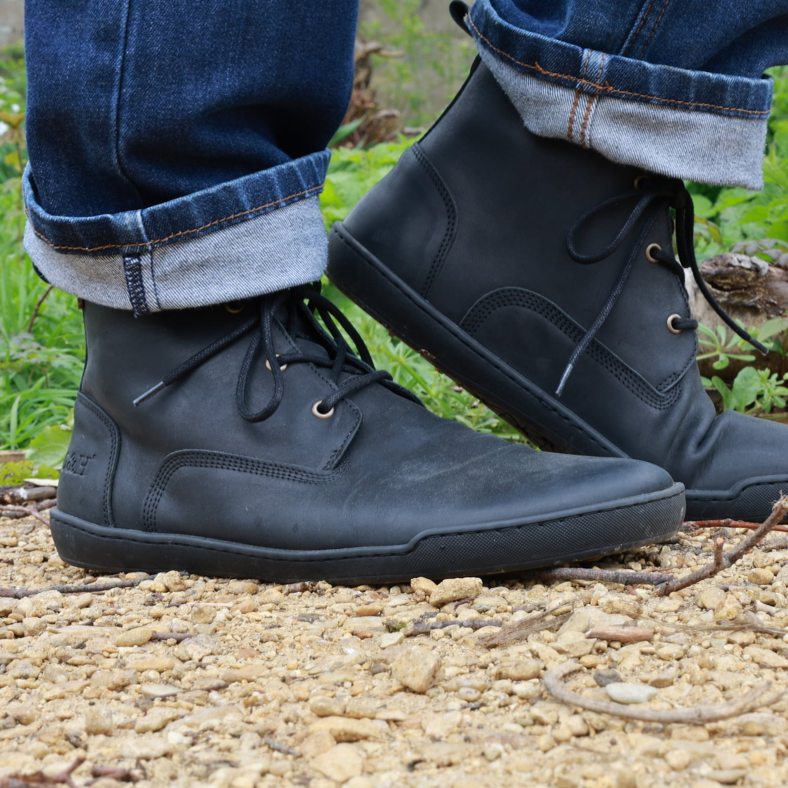 person wearing spruce barefoot chukka boots in black with blue jeans standing on gravel path