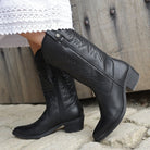 model wearing black leather texan hi cowboy boots with a white dress on a stone floor